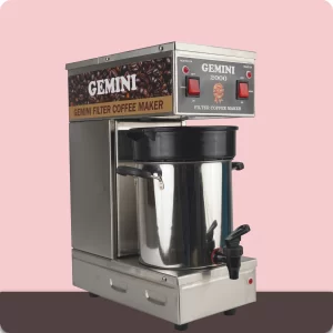 A stainless steel Gemini 2000 filter coffee maker with a black lid, red power switches, and a front tap, placed against a pink and brown background.